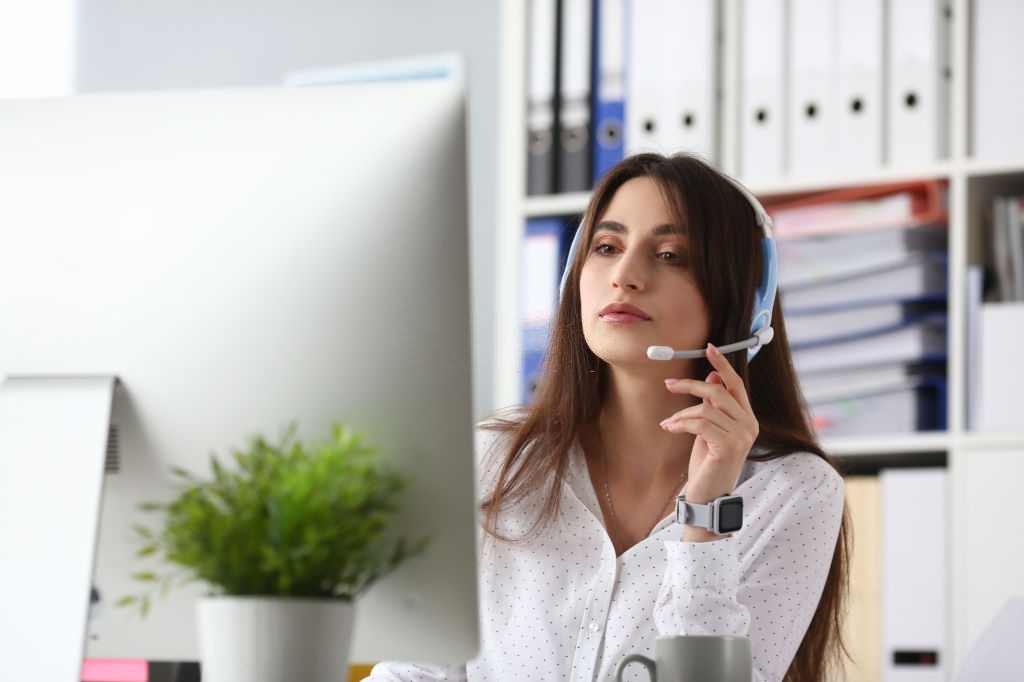 Portrait of lovely young woman wearing headset and answering questions on computer. Call center operator working in office. Folders on background. Telemarketing concept
