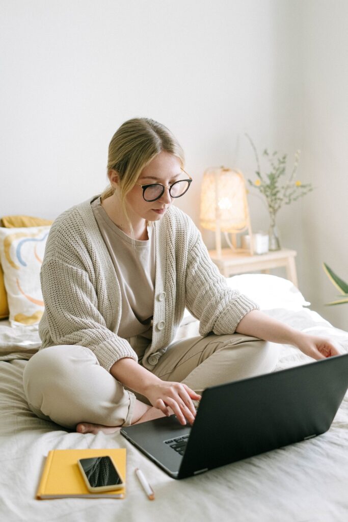 A woman sitting on her bed, with a notebook and a mobile phone next to her, browsing on a laptop