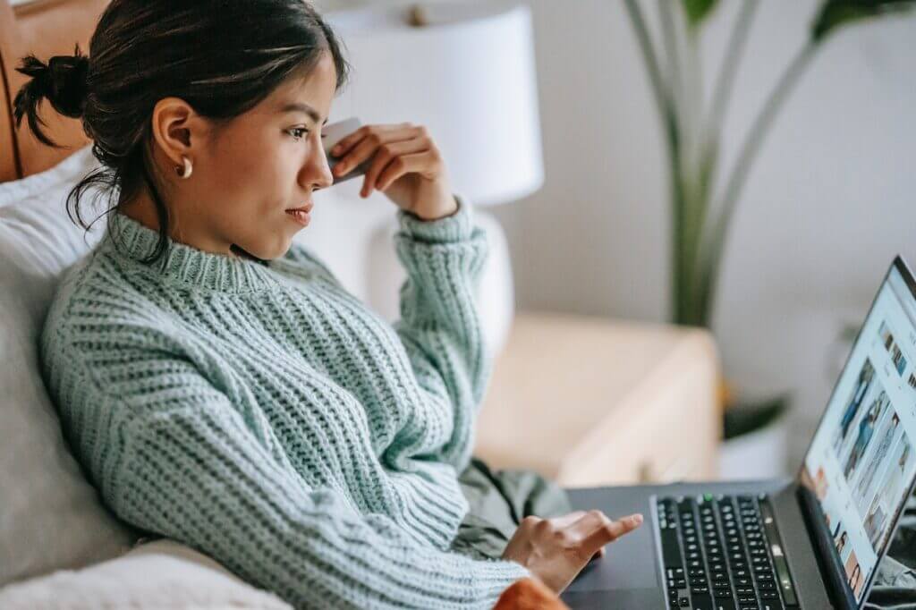 A woman on a couch, holding a credit card and browsing e-commerce site on the lap top in her lap