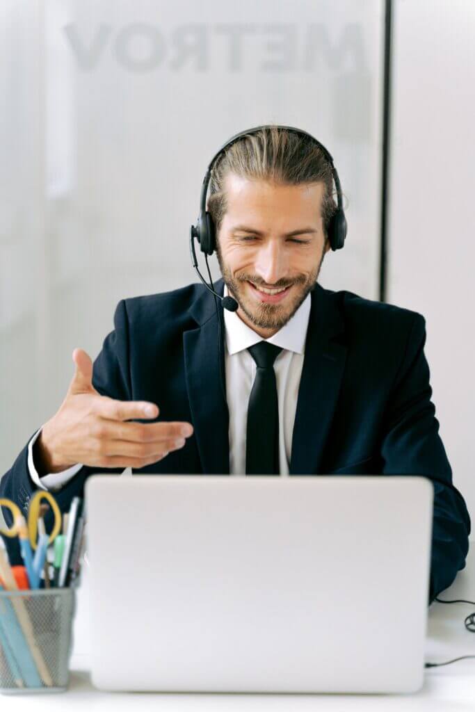 Man in a suit with a headset, looking at a laptop and gesticulating with his hand while smiling.