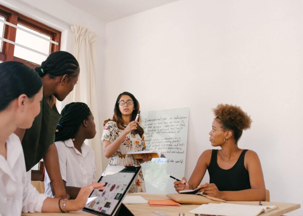 A woman presenting and writing on a whiteboard with four attentive female colleagues listening in an office environment.