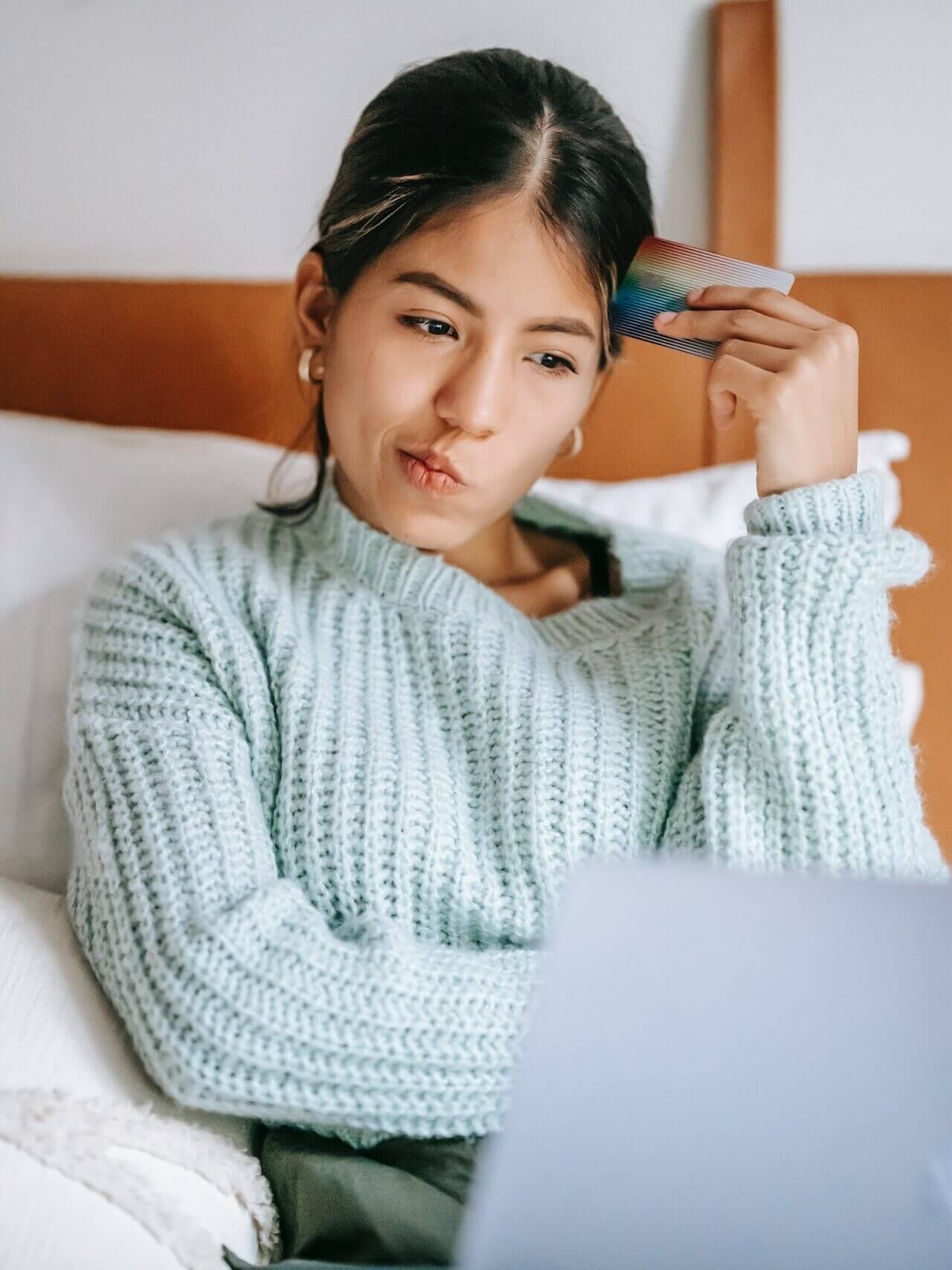 A woman with a laptop in front of her, laying in a bed, with a credit card in her hand