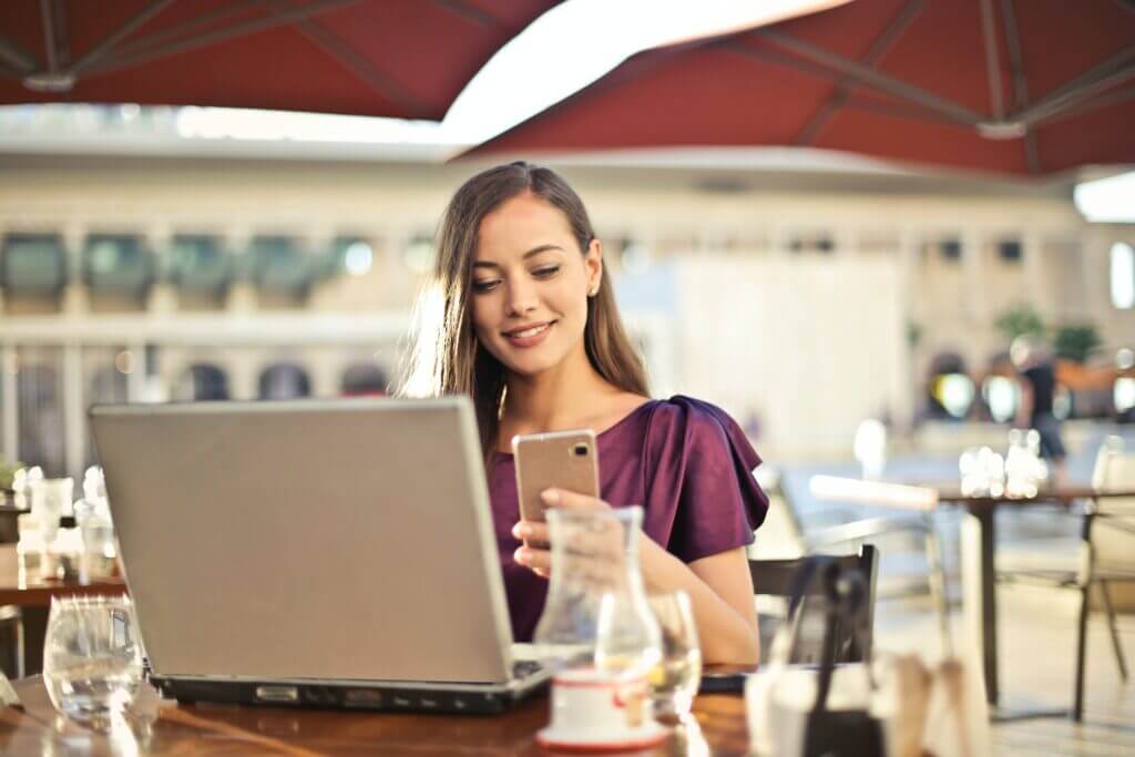 A girl with a lap top in front of her, looking at her phone, in an open cafe.