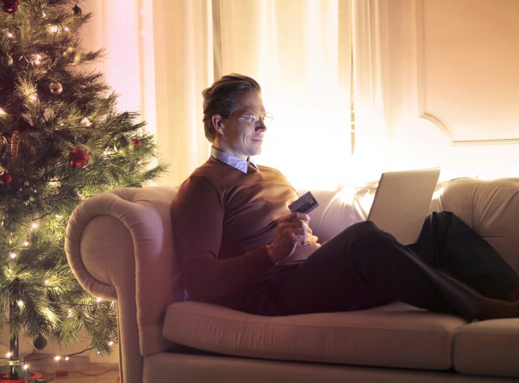 Man lying on a couch next to a Christmas tree, holding a credit card and laptop.