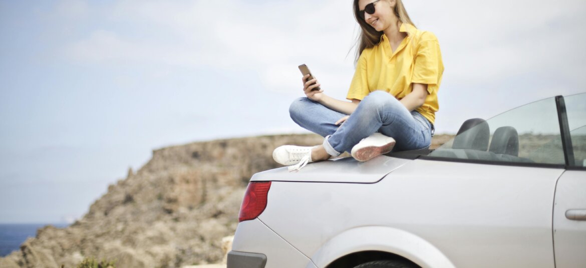 Woman in Yellow Blouse and Blue Jeans Taking Selfie While Sitting on Car