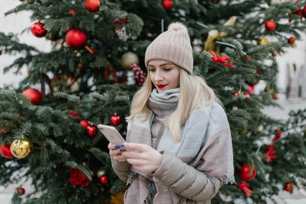 Blonde woman in winter clothing uses smartphone near decorated Christmas tree outdoors.