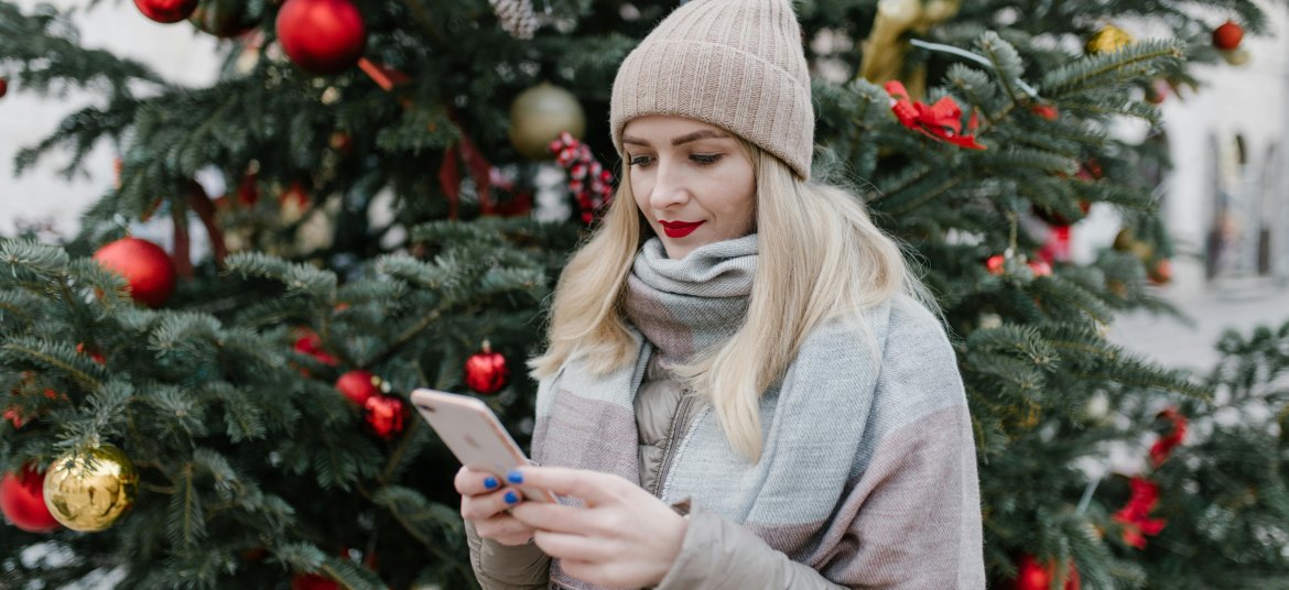 Blonde woman in winter clothing uses smartphone near decorated Christmas tree outdoors.